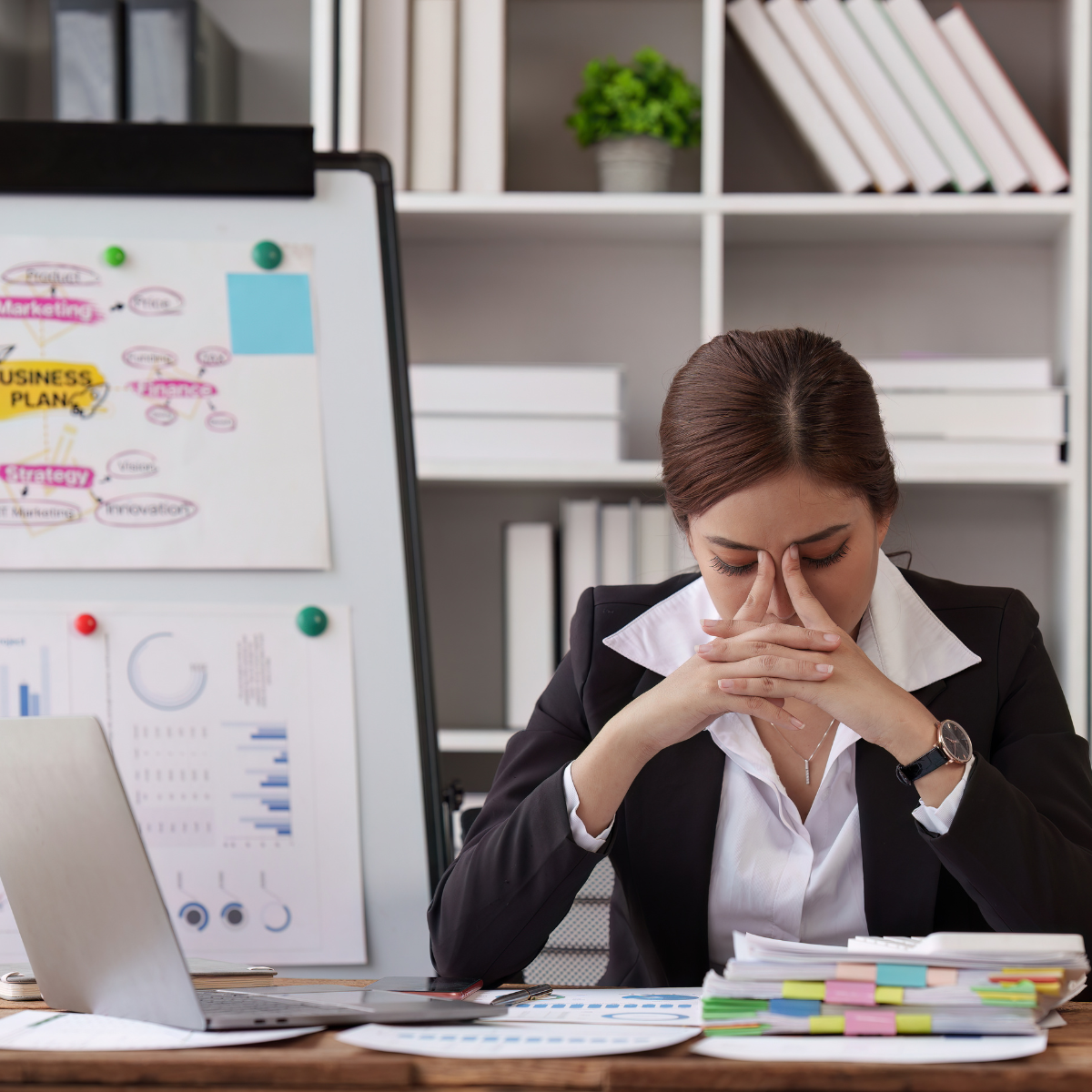 Woman in a business suit at a cluttered desk, hands pressed to her face, with a business plan diagram on the whiteboard behind her.